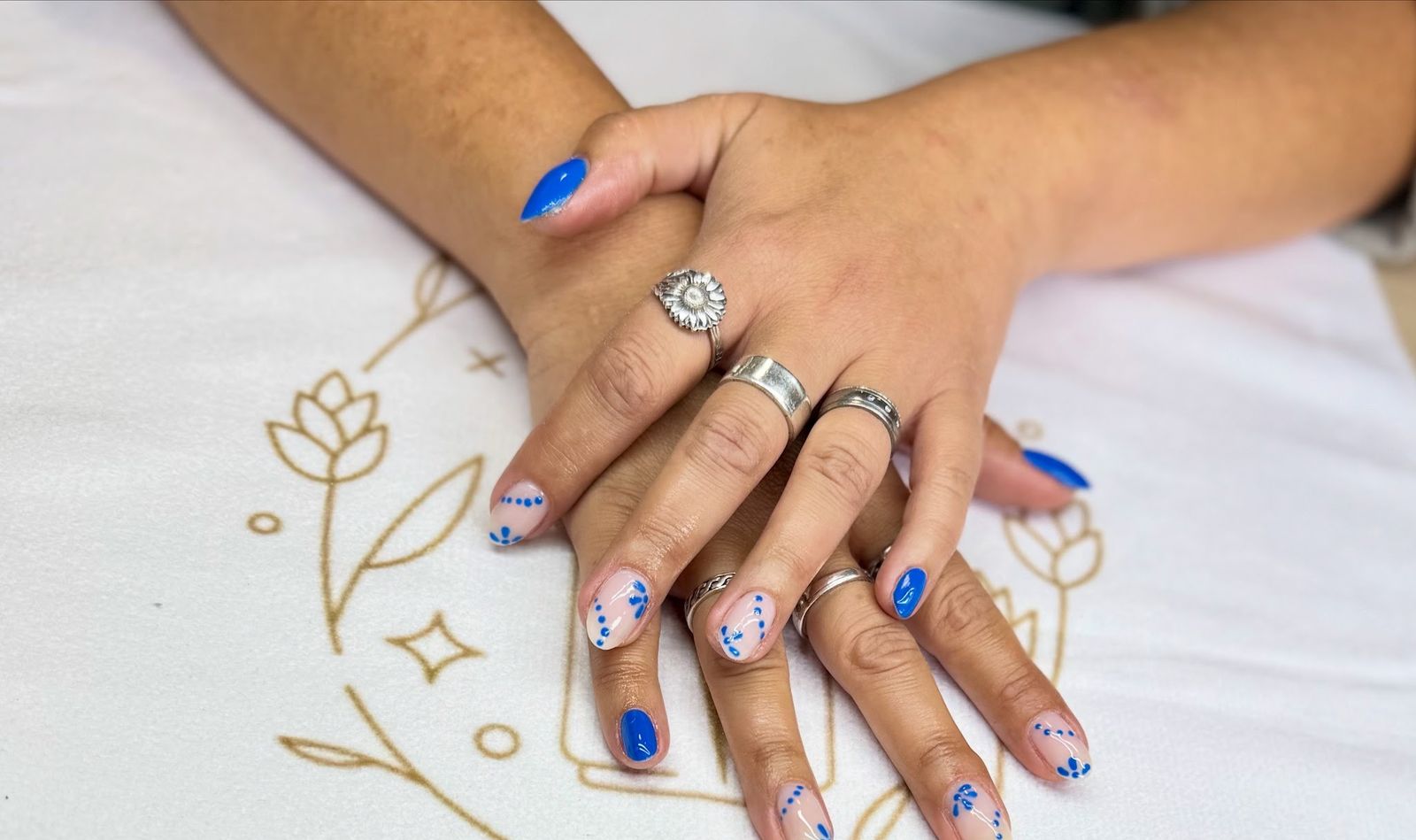 Korte nagels met blauwe stippenbloemetjes en kobaltblauwe accentnagels, op een doek met het gouden logo van de studio