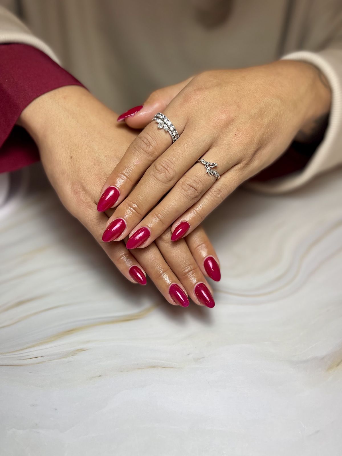 Twee handen met glanzende framboosrode amandelnagels en zilveren ringen, op een marmeren ondergrond