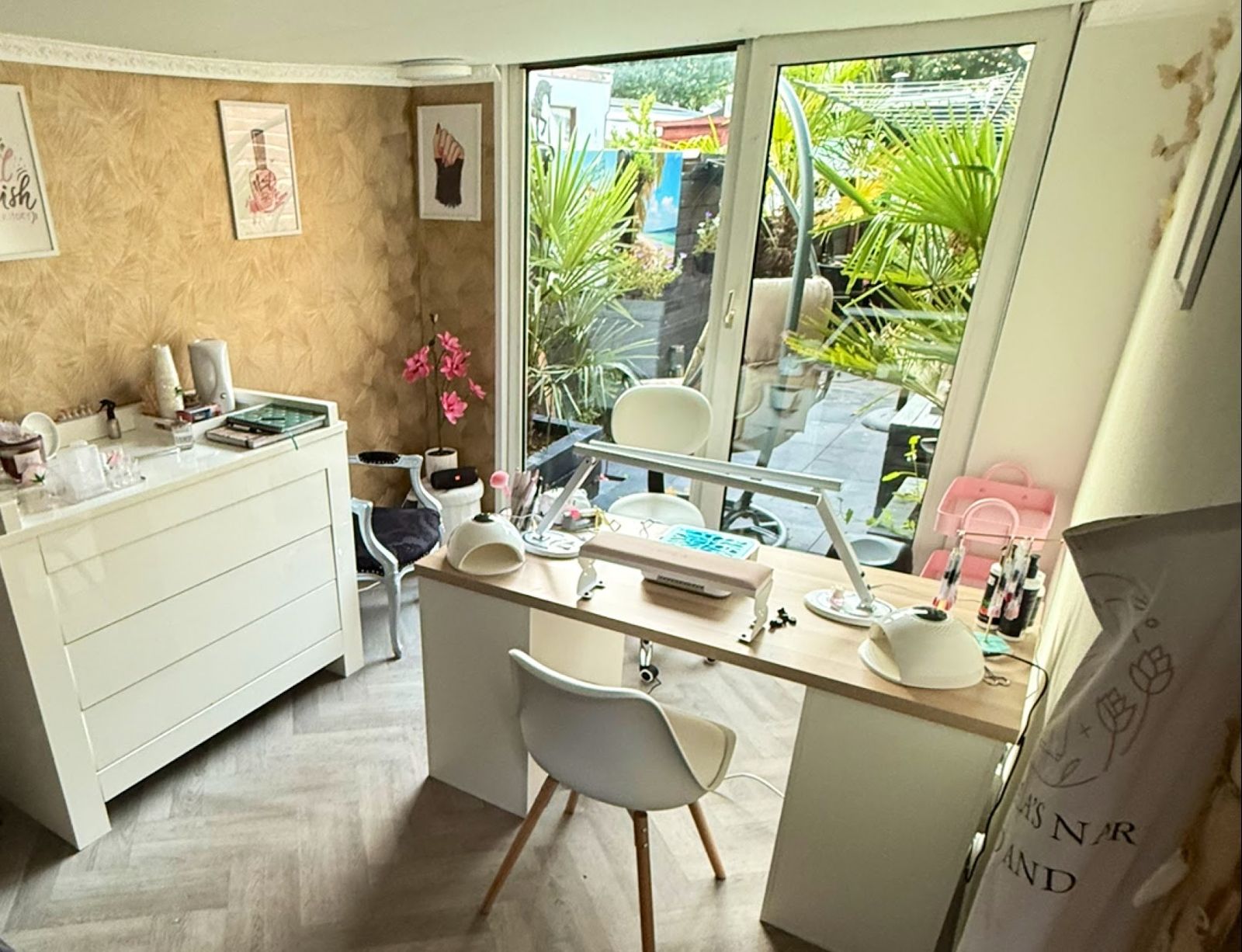De studio: een witte nageltafel met lampen, goudkleurig behang met palmbladeren, een witte ladekast en glazen schuifdeuren naar een groene tuin